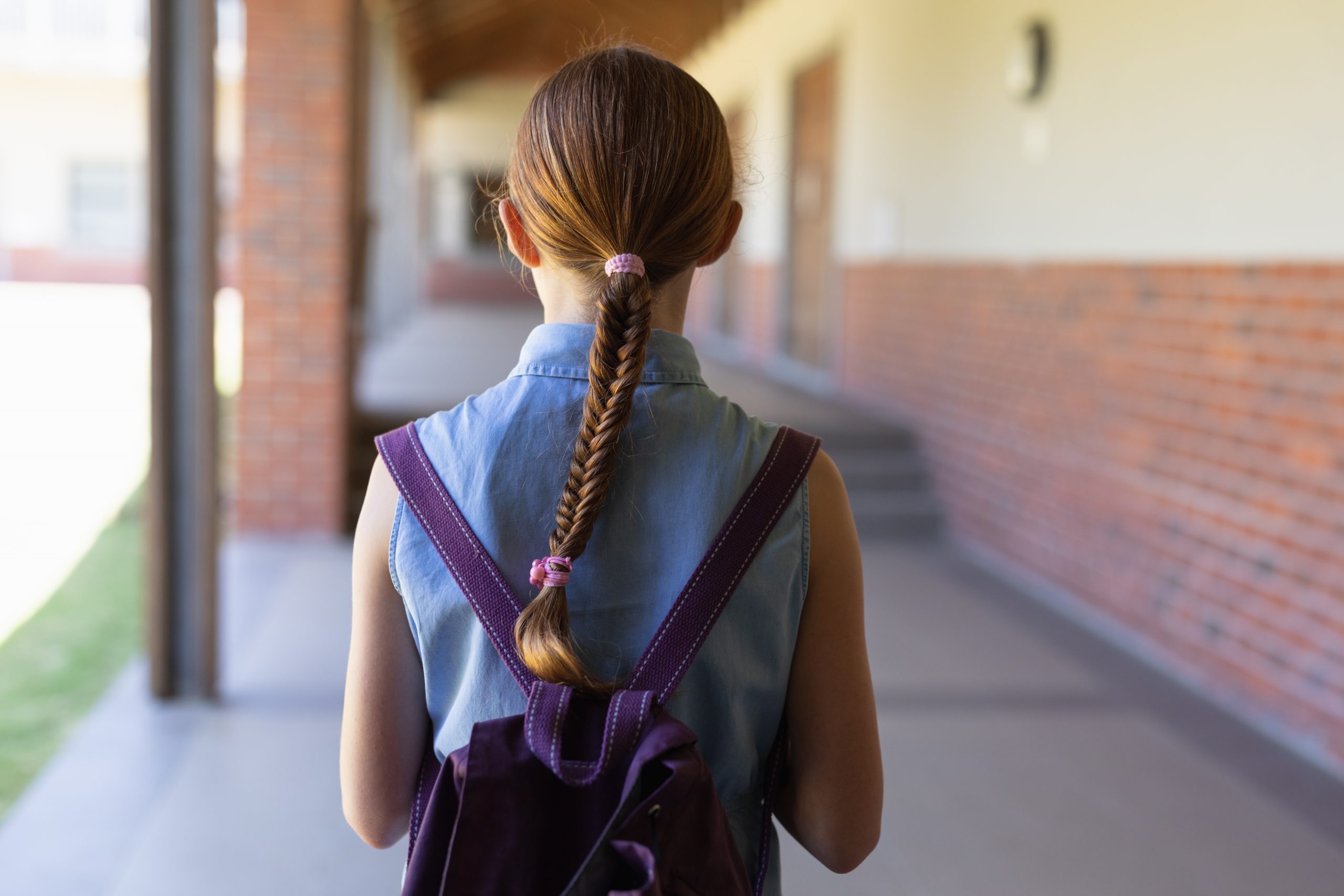 Front view of a Caucasian schoolgirl with red hair in a plait wearing a blue dress and a rucksack standing in the schoolyard at elementary school